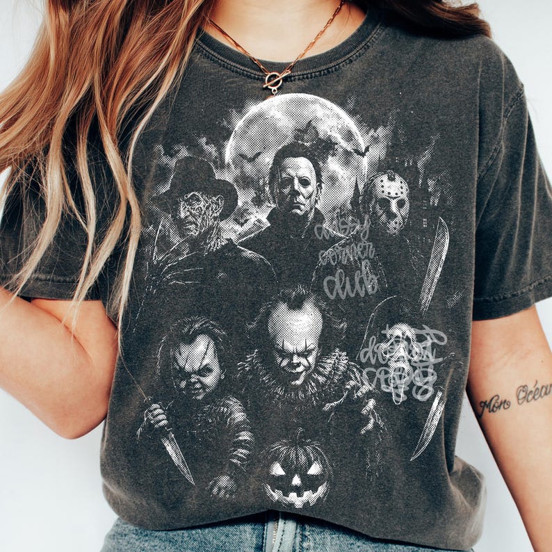 May include: A charcoal gray t-shirt featuring a white graphic print of five iconic horror movie characters, including a masked slasher, a clown, and a doll, set against a full moon background with bats and a jack-o'-lantern at the bottom.