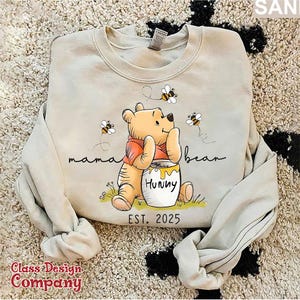 Może przedstawiać: Jasnobeżowa bluza z nadrukiem kresk&oacute;wkowego obrazka Winnie the Pooh trzymającego słoik miodu. Na bluzie nadrukowany jest tekst "mama bear" i "Hunny". Pod obrazkiem nadrukowany jest tekst "EST. 2025".