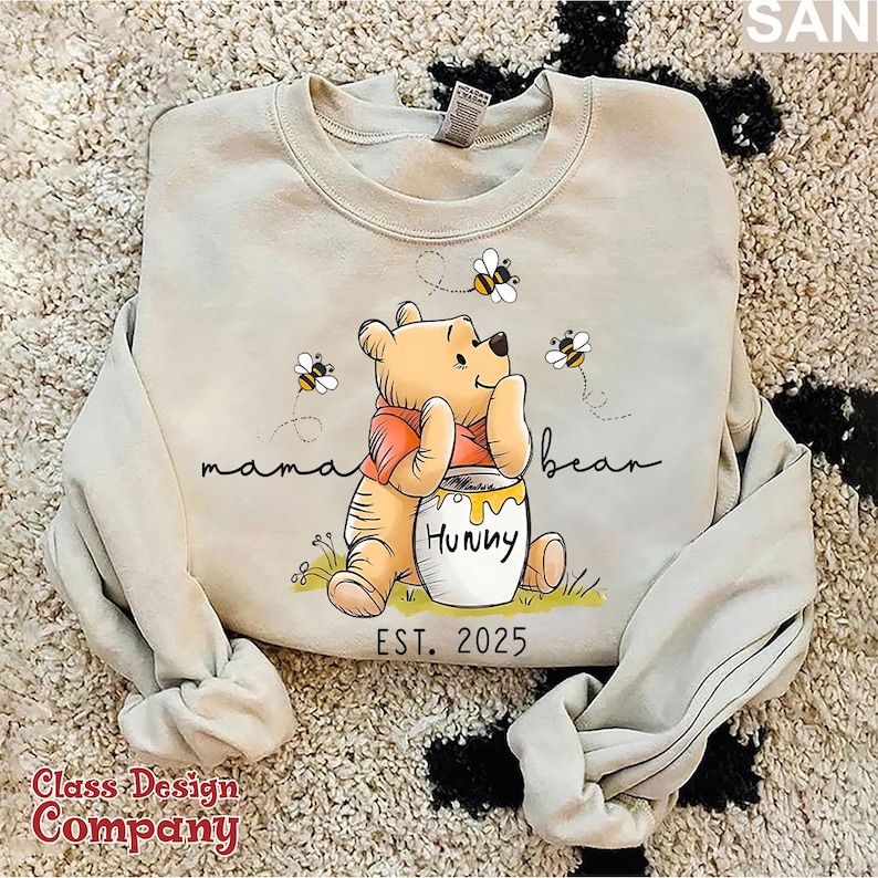 Może przedstawiać: Jasnobeżowa bluza z nadrukiem kresk&oacute;wkowego obrazka Winnie the Pooh trzymającego słoik miodu. Na bluzie nadrukowany jest tekst "mama bear" i "Hunny". Pod obrazkiem nadrukowany jest tekst "EST. 2025".