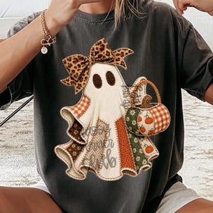 May include: A charcoal grey t-shirt featuring a central graphic of a ghost wearing a leopard print bow. The ghost is composed of various fabric-like patches in orange, green, and brown patterns, and it carries a small pumpkin-shaped basket.