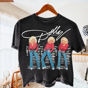 May include: A charcoal gray short-sleeve t-shirt featuring a white graphic print of Dolly Parton in three different poses. The singer wears a red polka-dot shirt and blue jeans. The text Dolly Parton is written in a white script font above.