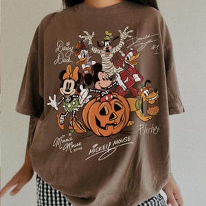 K&ouml;nnte beinhalten: Ein braunes Kurzarm-T-Shirt mit einem Grafikdruck von Disney-Figuren in Halloween-Kost&uuml;men, darunter Mickey Mouse in einem K&uuml;rbis, Minnie Mouse, Goofy, Donald Duck und Pluto, mit handgeschriebenen Namen und Unterschriften.
