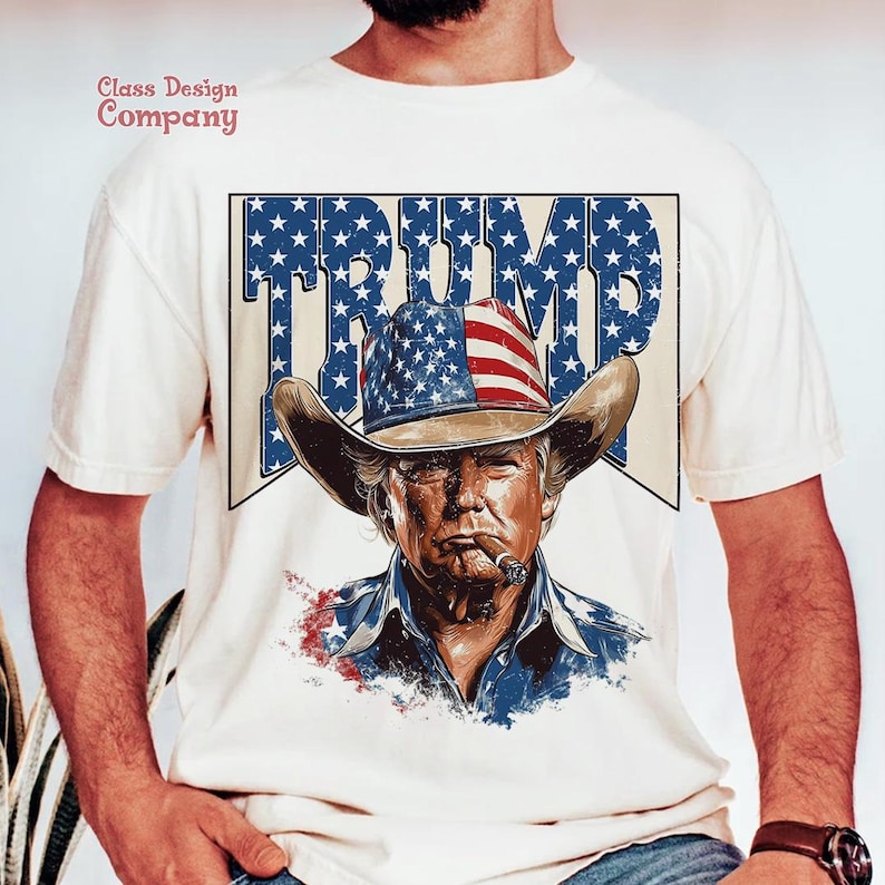 May include: White t-shirt with a graphic of a man wearing a cowboy hat with an American flag design. The text "TRUMP" is printed in large letters above the graphic. The text "Class Design Company" is printed in the top left corner of the shirt.