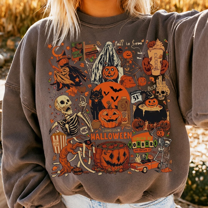 May include: A charcoal gray crewneck sweatshirt featuring a vibrant Halloween-themed graphic print. The design includes a skeleton, a black cat, pumpkins, a ghost, a cauldron, and an orange car, with text reading Fall is here and Long Live Halloween.