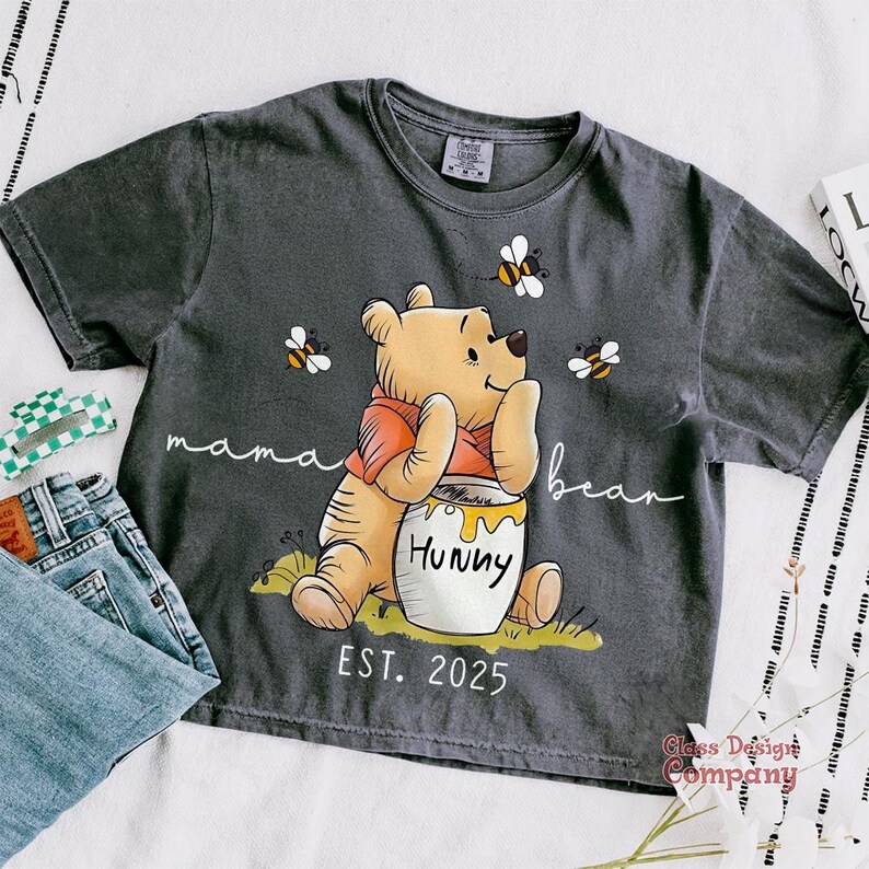Może przedstawiać: Szary T-shirt z kresk&oacute;wkowym wizerunkiem Kubusia Puchatka trzymającego słoik miodu. Na koszulce nadrukowany jest tekst "mama bear" i "hunny". Pod Kubusiem Puchatkiem nadrukowany jest tekst "EST. 2025".
