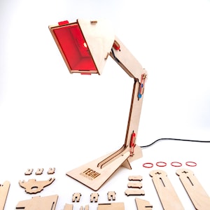 Könnte beinhalten: Ein Holz-Schreibtischlampen-Bausatz mit rot getöntem Licht. Die Lampe besteht aus hellem Holz und verfügt über verstellbare Gelenke. Auf dem Lampensockel steht der Text "TECHNI EDUCATION". Mehrere unmontierte Holzteile und rote Gummibänder sind enthalten.