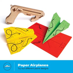 Op de afbeelding: Een houten papieren vliegtuigwerper met drie kleurrijke papieren vliegtuigen. De vliegtuigen zijn geel, groen en rood, met zwarte decoratieve lijnen. De tekst "Paper Airplanes" en "Try out different plane designs" staat onderaan.