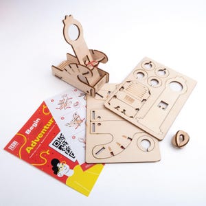 Könnte beinhalten: Ein Holzmodellbausatz einer Katapult, mit mehreren lasergeschnittenen Holzteilen, einer kleinen Holzkugel und einem Anleitungsblatt. Das Kit enthält den Text "Begin Your Adventure" auf dem Anleitungsblatt.