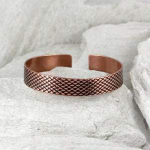 May include: A copper cuff bracelet with a textured, scale-like pattern. The open-ended bracelet is displayed on a light grey stone surface, showcasing its warm, reddish-brown colour and intricate design. This piece is a stylish accessory.