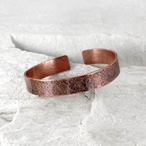 Op de afbeelding: Een koperen manchetarmband met een getextureerd oppervlak. De open armband heeft een rustieke, gehamerde uitstraling. Het warme, roodbruine metaal wordt gepresenteerd tegen een lichtgrijze achtergrond.