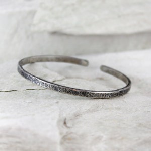 Op de afbeelding: Een zilveren manchetarmband met een getextureerd oppervlak. De open armband heeft een eenvoudig, minimalistisch ontwerp. Het metaal heeft een donkere, verouderde uitstraling, wat wijst op een vintage of handgemaakte stijl. Dit sieraad is geschikt voor dagelijks gebruik.
