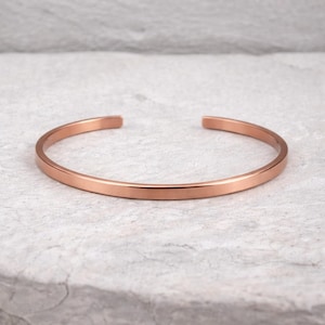 May include: A rose gold cuff bracelet. The open-ended bracelet has a simple, minimalist design. The metal has a smooth, polished finish. The bracelet is shown on a light gray surface.