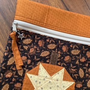 May include: A fabric zipper pouch featuring a dark brown floral pattern with orange accents. The top section is solid orange fabric. A white zipper closure includes a silver star-shaped charm and an orange fabric pull tab. A quilted star design.