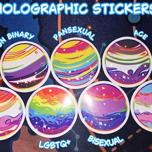 May include: Holographic stickers with colorful planet designs representing LGBTQ+ identities. Each sticker features a unique color scheme and text labels such as "Non Binary," "Pansexual," "Ace," "Trans," "Lesbian," "LGBTQ*," and "Bisexual."
