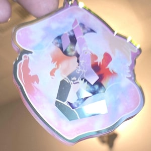 以下が含まれることがあります： ピンク、紫、オレンジの色合いのシルエットが、銀河のような背景に重ねられたホログラフィックチャーム。チャームはシルバーの縁取りで、上部に小さなループが付いています。