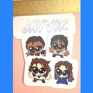 以下が含まれることがあります： 大きな目と様々な髪型をした、4つの漫画のキャラクターのステッカー。ステッカーはピンク色の表面にあり、背景には定規があります。「JAYVIK」という言葉がパステルカラーで上部に書かれています。