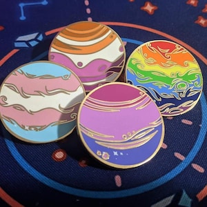 May include: Four circular enamel pins with planet designs, each displaying a different colour scheme. The pins represent various LGBTQ+ pride flags, including the transgender, lesbian, gay, and bisexual flags. The pins have gold borders and are set against a dark blue background.