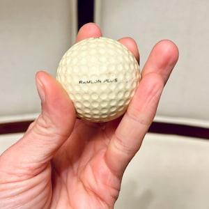 K&ouml;nnte beinhalten: Ein cremefarbener Golfball mit einer geriffelten Oberfl&auml;che. Der Text "RAMLON PLUS" ist in Schwarz auf den Ball gedruckt. Der Ball wird in der Hand einer Person gehalten.