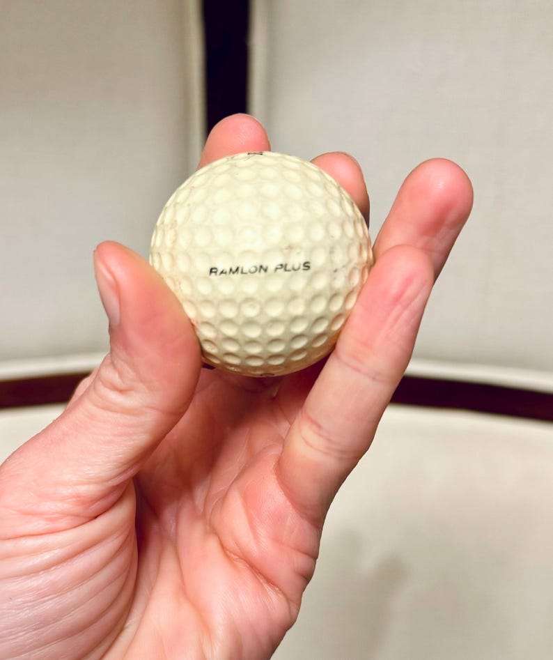 K&ouml;nnte beinhalten: Ein cremefarbener Golfball mit einer geriffelten Oberfl&auml;che. Der Text "RAMLON PLUS" ist in Schwarz auf den Ball gedruckt. Der Ball wird in der Hand einer Person gehalten.