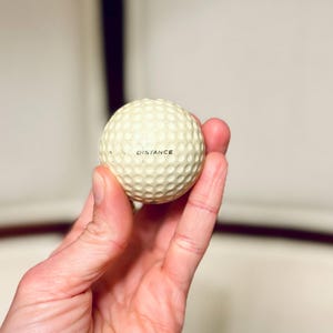 K&ouml;nnte beinhalten: Ein Golfball mit einer genoppten Oberfl&auml;che wird in einer Hand gehalten. Das Wort "DISTANCE" ist schwarz auf dem Ball aufgedruckt. Der Ball ist cremefarben und scheint neu zu sein.