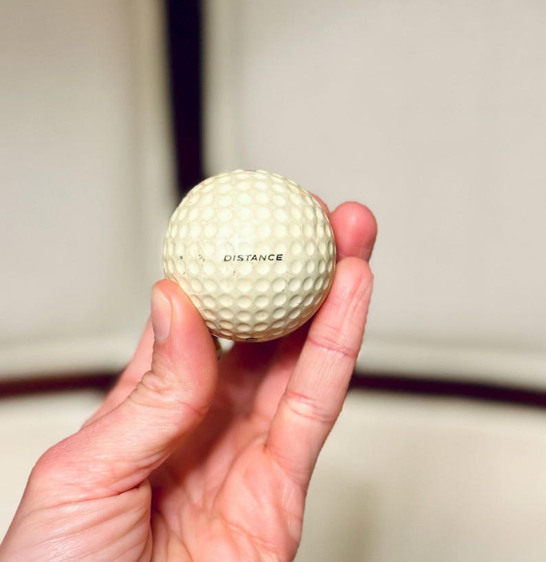 K&ouml;nnte beinhalten: Ein Golfball mit einer genoppten Oberfl&auml;che wird in einer Hand gehalten. Das Wort "DISTANCE" ist schwarz auf dem Ball aufgedruckt. Der Ball ist cremefarben und scheint neu zu sein.