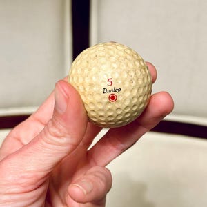 K&ouml;nnte beinhalten: Ein Vintage-Golfball mit einer genoppten Oberfl&auml;che. Der cremefarbene Ball hat die Nummer "5", das Wort "Dunlop" und einen roten Kreis aufgedruckt. Der Ball weist Gebrauchsspuren auf. Er wird in einer Hand gehalten.