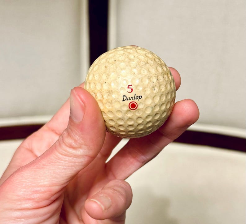 K&ouml;nnte beinhalten: Ein Vintage-Golfball mit einer genoppten Oberfl&auml;che. Der cremefarbene Ball hat die Nummer "5", das Wort "Dunlop" und einen roten Kreis aufgedruckt. Der Ball weist Gebrauchsspuren auf. Er wird in einer Hand gehalten.