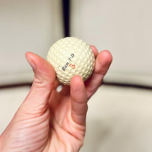 K&ouml;nnte beinhalten: Ein cremefarbener Golfball mit dem Text "Ram 3-D" und der roten Zahl "3". Der Ball hat eine strukturierte Oberfl&auml;che. Der Golfball wird in einer Hand gehalten.