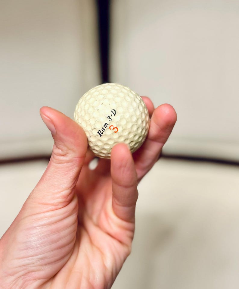 K&ouml;nnte beinhalten: Ein cremefarbener Golfball mit dem Text "Ram 3-D" und der roten Zahl "3". Der Ball hat eine strukturierte Oberfl&auml;che. Der Golfball wird in einer Hand gehalten.