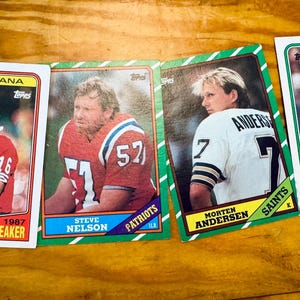 Pode incluir: Quatro cartões colecionáveis de futebol americano Topps retangulares apresentando Joe Montana com camisa vermelha, Steve Nelson com camisa vermelha dos Patriots, Morten Andersen com camisa branca dos Saints e Harry Hamilton com camisa verde.