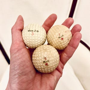 K&ouml;nnte beinhalten: Drei Vintage-Golfb&auml;lle mit genoppter Oberfl&auml;che werden in einer Hand gehalten. Die Golfb&auml;lle sind cremefarben mit schwarzen und roten Markierungen. Auf einem Ball steht "Ram 3-D" mit der Nummer "3". Die anderen beiden B&auml;lle tragen den Namen "Muscle" und die Nummern "2" und "5".