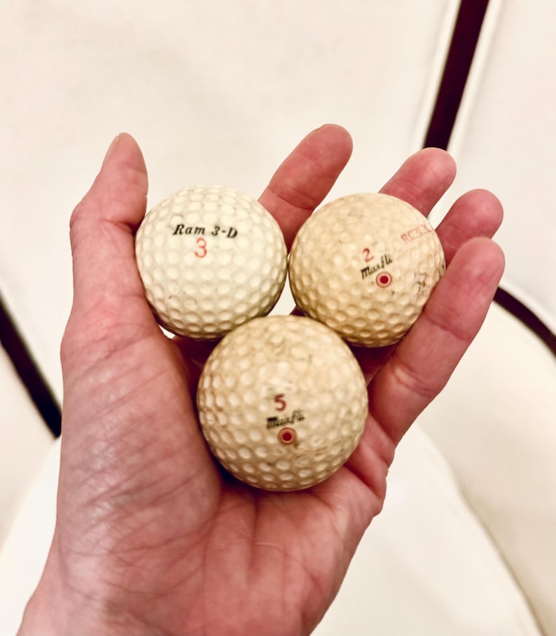 K&ouml;nnte beinhalten: Drei Vintage-Golfb&auml;lle mit genoppter Oberfl&auml;che werden in einer Hand gehalten. Die Golfb&auml;lle sind cremefarben mit schwarzen und roten Markierungen. Auf einem Ball steht "Ram 3-D" mit der Nummer "3". Die anderen beiden B&auml;lle tragen den Namen "Muscle" und die Nummern "2" und "5".