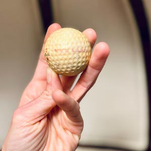 K&ouml;nnte beinhalten: Ein gebrauchter Golfball mit einer geriffelten Oberfl&auml;che, gehalten in einer Hand. Der Ball ist cremefarben mit rotem Text. Der Golfball hat einen Durchmesser von etwa 4,27 cm.