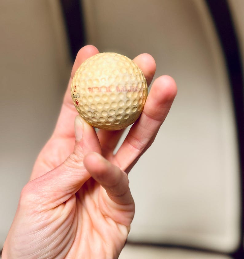 K&ouml;nnte beinhalten: Ein gebrauchter Golfball mit einer geriffelten Oberfl&auml;che, gehalten in einer Hand. Der Ball ist cremefarben mit rotem Text. Der Golfball hat einen Durchmesser von etwa 4,27 cm.