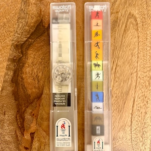 Puede incluir: Dos relojes Swatch Quartz en estuches de plástico transparente. Un reloj tiene una correa blanca y una esfera plateada. El otro estuche contiene bloques coloridos con temática deportiva. Ambos estuches tienen el logotipo de Swatch y texto de los Juegos Olímpicos de 1996.