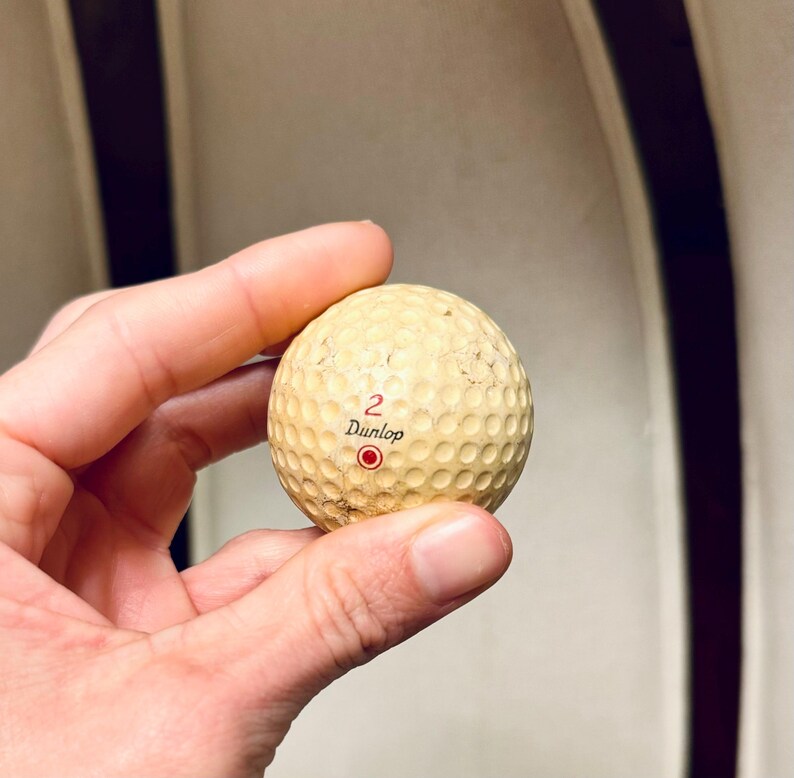 K&ouml;nnte beinhalten: Ein gebrauchter Dunlop Golfball, cremefarben, mit der Nummer "2" und dem Markennamen in Schwarz aufgedruckt. Unter der Nummer befindet sich ein roter Kreis. Der Ball hat eine strukturierte Oberfl&auml;che.
