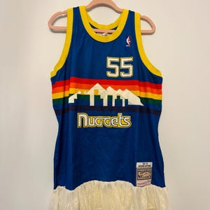 Può includere: Una maglia da basket blu dei Denver Nuggets con il numero 55 in bianco. La maglia presenta un design a strisce arcobaleno e il nome della squadra. Un bordo in pizzo color crema è aggiunto sul fondo. La maglia è appesa a un gancio rosa.