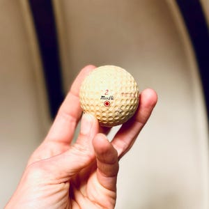 K&ouml;nnte beinhalten: Ein cremefarbener Golfball mit einer geriffelten Oberfl&auml;che. Die Zahl "2" und der Markenname "Maxfli" sind rot aufgedruckt. Ein roter Kreis ist ebenfalls sichtbar. Der Ball wird in einer Hand gehalten.