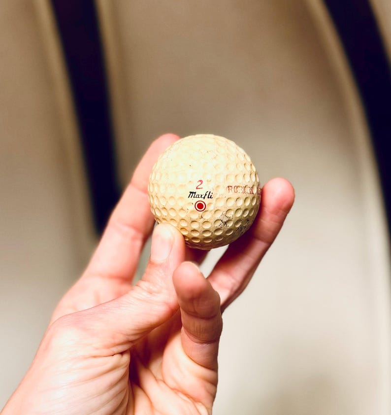 K&ouml;nnte beinhalten: Ein cremefarbener Golfball mit einer geriffelten Oberfl&auml;che. Die Zahl "2" und der Markenname "Maxfli" sind rot aufgedruckt. Ein roter Kreis ist ebenfalls sichtbar. Der Ball wird in einer Hand gehalten.