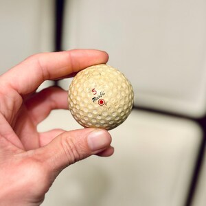 K&ouml;nnte beinhalten: Ein Vintage-Golfball mit einer genoppten Oberfl&auml;che. Der Ball ist cremefarben mit der Zahl "5" und dem Wort "Maxfli" in Schwarz aufgedruckt, darunter ein roter Kreis. Der Ball weist Gebrauchsspuren auf. Der Ball wird in einer Hand gehalten.