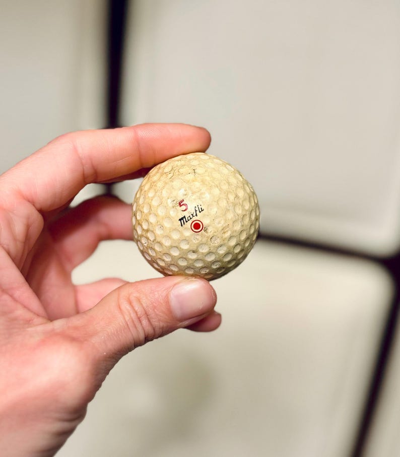K&ouml;nnte beinhalten: Ein Vintage-Golfball mit einer genoppten Oberfl&auml;che. Der Ball ist cremefarben mit der Zahl "5" und dem Wort "Maxfli" in Schwarz aufgedruckt, darunter ein roter Kreis. Der Ball weist Gebrauchsspuren auf. Der Ball wird in einer Hand gehalten.