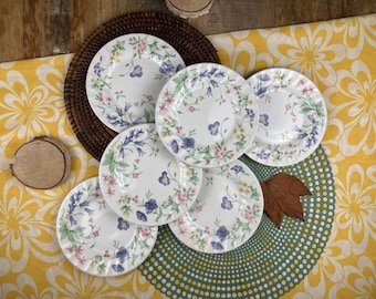 May include: Six white ceramic plates with a floral design in shades of pink, purple, and green. The plates have a scalloped edge and are arranged on a yellow and green patterned surface.