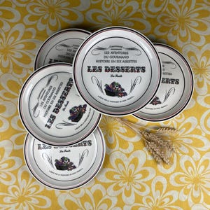 May include: A collection of white ceramic dessert plates with black and red rims. Each plate is inscribed with "LES DESSERTS" and a fruit basket graphic. The plates are displayed on a yellow floral tablecloth.
