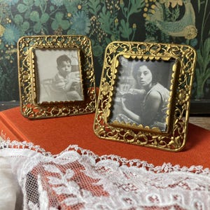 Può includere: Due cornici dorate ornate con foto in bianco e nero all'interno. Le cornici sono rettangolari con intricati disegni floreali e a volute. Sono appoggiate su un libro rosso, con pizzo bianco in primo piano.