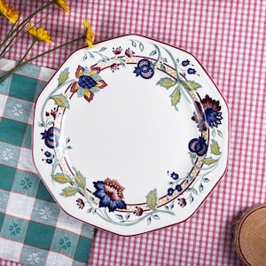 Puede incluir: Plato octogonal blanco con un diseño floral con flores azules, amarillas y burdeos. El plato tiene un borde burdeos y está sobre un mantel a cuadros rojo y blanco. Vajilla decorativa.