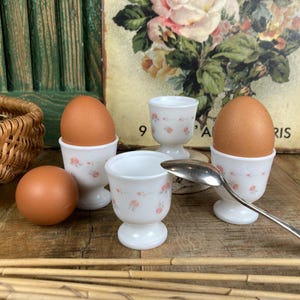 May include: A still life featuring brown eggs and white egg cups with pink floral designs. A silver spoon rests near the cups. The background includes a vintage floral print and a wooden surface.