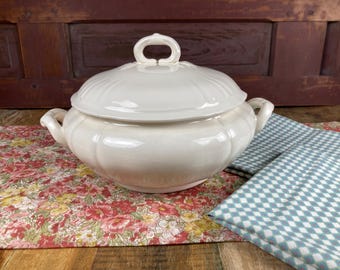 May include: A white ceramic serving dish with a lid and two handles. The dish has a fluted design and is sitting on a floral patterned table runner.