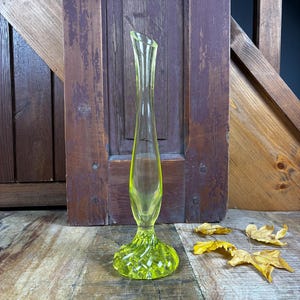 May include: A slender, yellow-green glass vase with a flared, textured base and a gently curved top. The vase is approximately 10 inches tall, showcasing a delicate, elongated design. The vase is set against a rustic wooden backdrop.