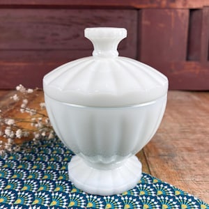 Puede incluir: Un cuenco blanco con tapa y diseño acanalado. El cuenco tiene una base de pedestal y un adorno decorativo en la tapa. El cuenco está hecho de vidrio opaco y se encuentra sobre una tela azul y amarilla estampada.