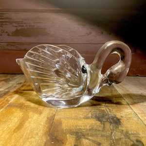 Peut inclure: Un plat en forme de cygne en verre transparent, avec un motif texturé en forme d'éventail sur le corps et une poignée incurvée en forme de cou. Le plat repose sur une surface en bois.